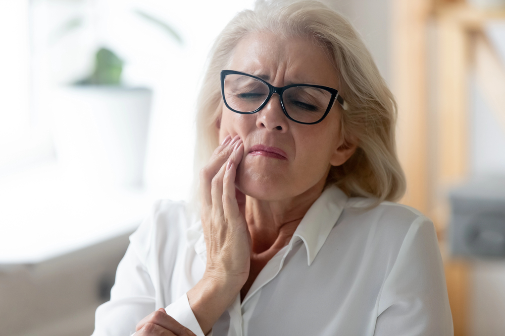 older woman with blonde hair, black glasses, and white shirt, wincing from TMJ pain